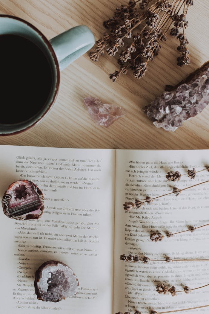 A relaxing flat lay scene with coffee, an open book, crystals, and dried flowers on a wooden table.